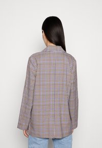 Žena v hnědo-bílo-modré kostkované blazeru s dlouhými rukávy, zobrazená zezadu, v kombinaci se světle modrými džíny na jednotném pozadí.