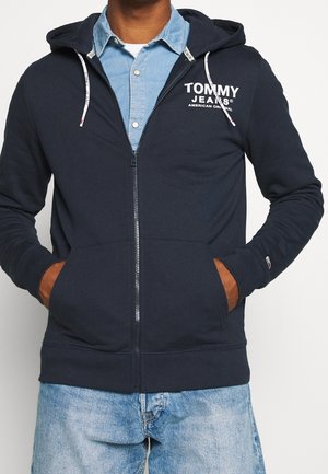 Sudadera con cremallera en color navy, con cordones grises y bolsillos frontales. Presenta el logo "TOMMY JEANS" en color blanco. Combinada con una camisa de mezclilla clara debajo.