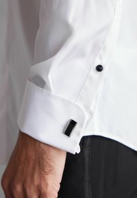 Manșeta unei cămăși albe dintr-un material neted, cu un strat interior texturat, având un buton negru și un buton de manșetă dreptunghiular în culorile negru și argintiu.