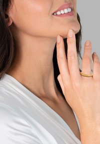 Zlat prstan z teksturiranim, zvitim dizajnom na prstu, ob kateri se jasno vidijo gladki toni kože in svetla barvna ozadja.