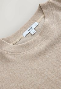 Gros plan sur le col d'un pull beige côtelé avec une étiquette en tissu blanche indiquant « NEXT » et « Made in China ».