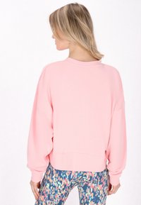 Felpa rosa chiaro con orlo corto, scollatura rotonda e vestibilità ampia. Polsini a coste e texture morbida; indossata con leggings a fantasia.
