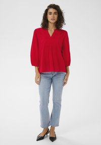 Blouse rouge avec un col en V et des détails plissés, associée à un jean évasé bleu clair et des talons noirs à bout pointu. Texture de tissu lisse.