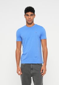 Μπλε βαμβακερό μπλουζάκι με στρογγυλή λαιμόκοψη, κοντά μανίκια και μια μικρή λεπτομέρεια λογότυπου στο στήθος. Το ύφασμα φαίνεται απαλό και ελαφρώς ραφανιστό.