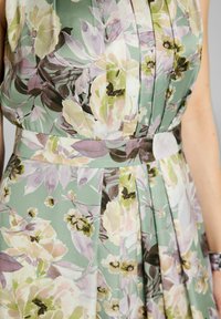 Robe fleurie avec un fond vert et un design plissé. Comprend de grandes fleurs et feuilles pastel dans des teintes de rose, de jaune et de lavande.