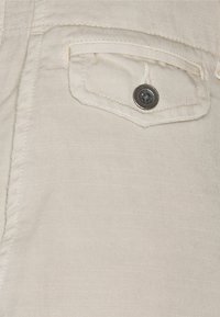 Detail béžové látky s klopovou kapsou zabezpečenou jedním tmavým knoflíkem na oděvu.