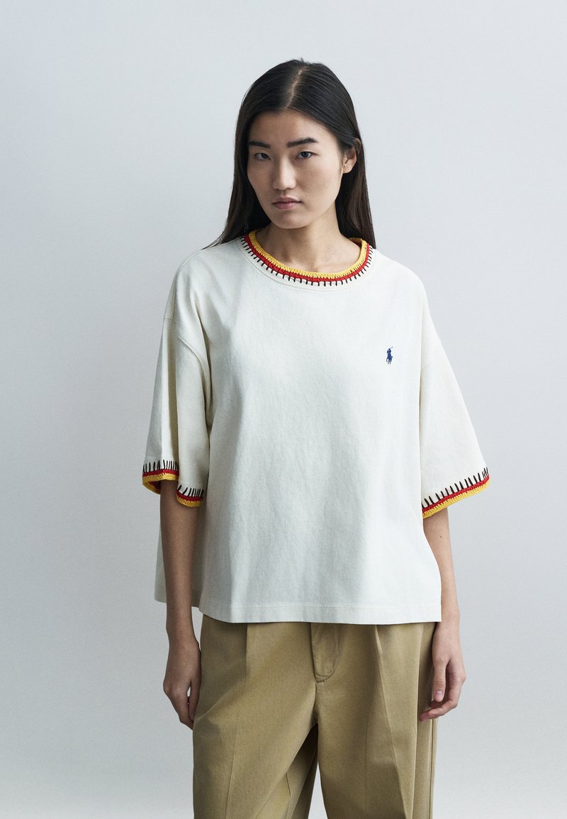 Biele bavlnené tričko s krátkymi rukávmi, s guľatým výstrihom ozdobeným červenou a žltou výšivkou a malým logom na hrudi.