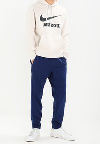 Ljusgrå huvtröja med svart "Just Do It"-text, kombinerad med marinblå joggingbyxor och vita sneakers. Avslappnad design, mjukt tyg, avslappnad passform.