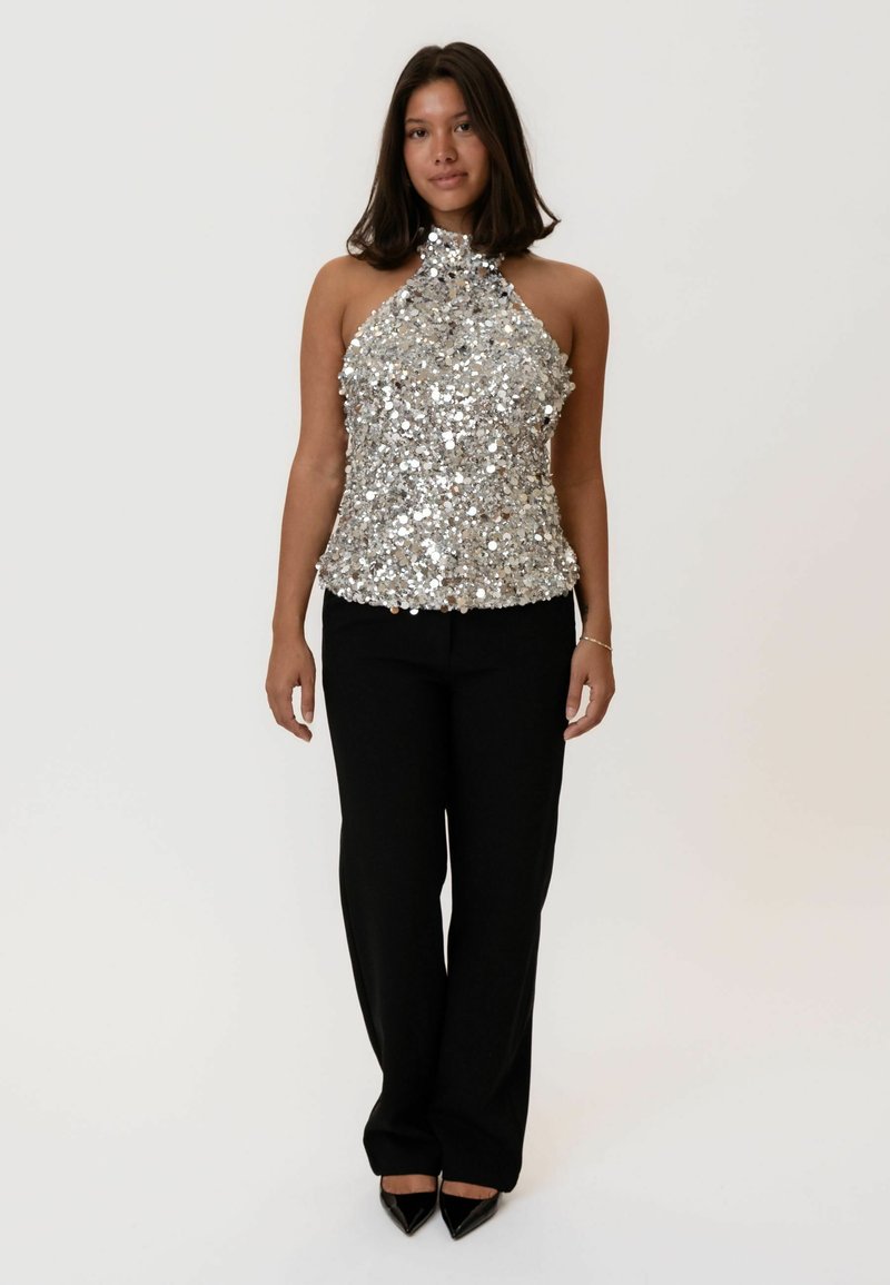 Top halter di paillettes argento abbinato a pantaloni neri su misura. Il top presenta un collo alto e una silhouette aderente.