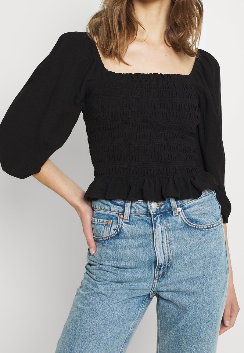 Blouse noire smockée avec manches bouffantes et ourlet à volants, associée à un jean taille haute bleu clair. Caractéristiques : encolure carrée et tissu texturé.