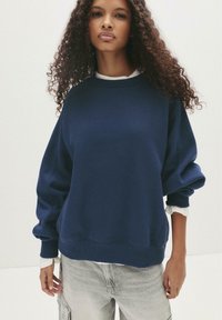 Felpa oversize blu navy con maniche lunghe, polsini a costine e una consistenza morbida. Indossata sopra una camicia bianca con colletto, caratterizzata da un collo rotondo.