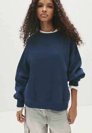 Felpa oversize blu navy con maniche lunghe, polsini a costine e una consistenza morbida. Indossata sopra una camicia bianca con colletto, caratterizzata da un collo rotondo.