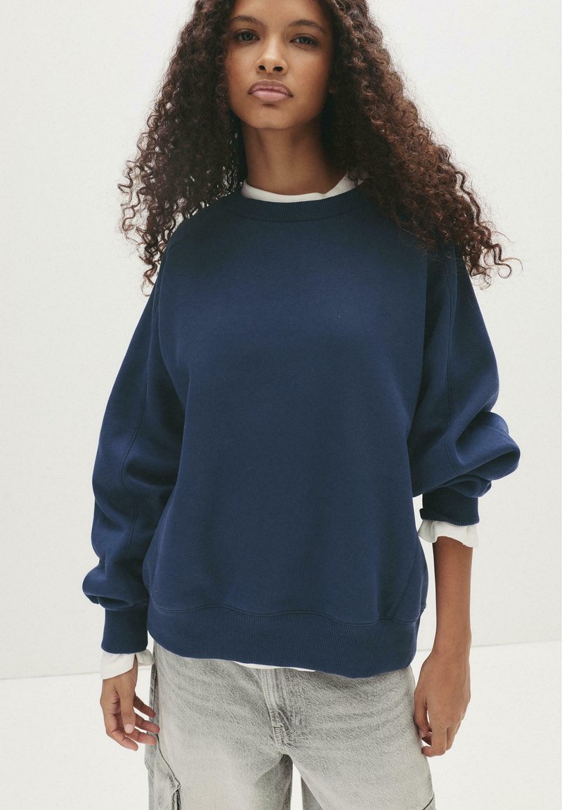 Felpa oversize blu navy con maniche lunghe, polsini a costine e una consistenza morbida. Indossata sopra una camicia bianca con colletto, caratterizzata da un collo rotondo.