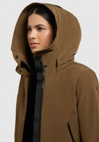 Veste à capuche marron avec fermeture zippée, présentant des accents noirs, des boutons-pression et des poches latérales. Tissu lisse avec une finition mate.