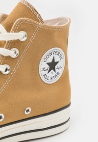 Zapatilla alta de lona color beige con una suela de goma blanca, que presenta ojales, cordones beige y un parche circular en blanco y negro con el logo.