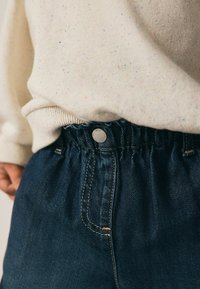 Temno modri traper hlače z nabrano elastiko in srebrnim gumbom. Mehak kremni pulover z večbarvnimi pikami je delno viden zgoraj.