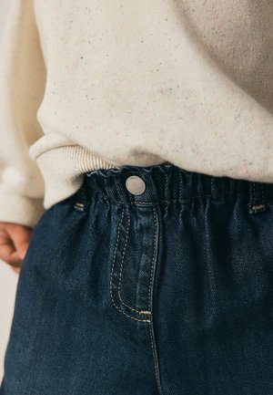 Temno modri traper hlače z nabrano elastiko in srebrnim gumbom. Mehak kremni pulover z večbarvnimi pikami je delno viden zgoraj.
