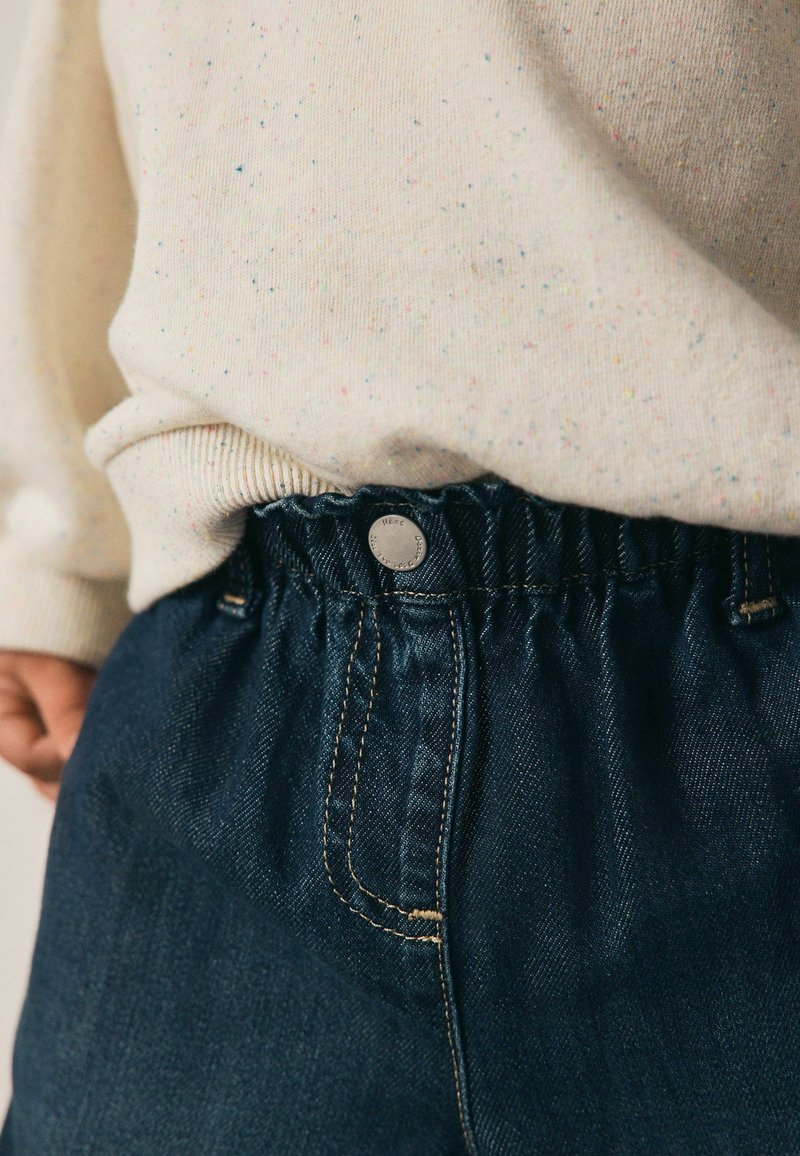 Temno modri traper hlače z nabrano elastiko in srebrnim gumbom. Mehak kremni pulover z večbarvnimi pikami je delno viden zgoraj.