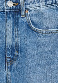 Gros plan d’un jean en denim bleu montrant la poche avant, la passants de ceinture, le bouton en métal et les coutures cousues avec un fil jaune.