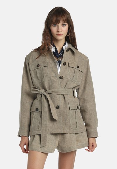 Femme portant une veste beige ceinturée avec quatre poches à boutons et un short assorti, accompagnés d'une chemise blanche et d'une écharpe à motifs.