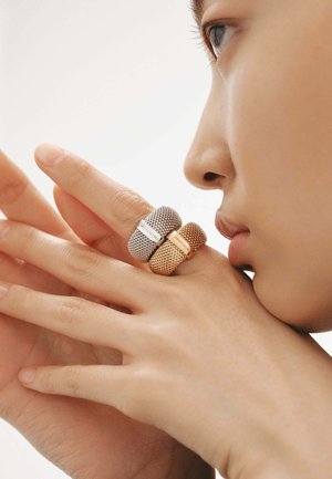 Tres anillos de malla en diferentes tonalidades: plateado con una banda blanca, dorado con un diseño texturizado. Se muestra una mano que los lleva puestos contra un fondo neutro.