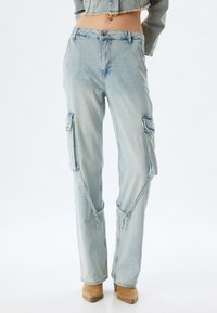 Pantalones cargo de denim azul claro con bolsillos con tapa, un ajuste relajado y cremalleras ajustables en los tobillos. Textura desgastada con un ligero desvanecimiento.