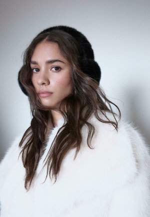 Black fur earmuffs, soft texture, worn with a white fur coat featuring a high collar. Long, wavy dark hair partially visible. Neutral background.