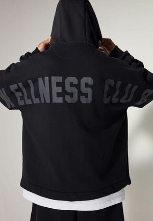 Black hooded sweatshirt featuring "WELLNESS CLUB" in large, bold grey letters across the back. Soft fabric with a relaxed fit.