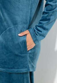 Prenda de forro polar turquesa con un bolsillo lateral y textura suave, con dobladillo redondeado y mangas largas. Primer plano de una mano en el bolsillo.