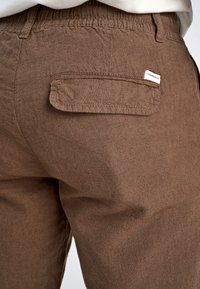 Brązowe lniane spodnie z teksturowanej tkaniny, z tylną kieszenią i białą metką. Pas jest elastyczny dla zapewnienia komfortu.