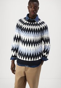Maglione a maglia decorato in bianco, blu navy e azzurro chiaro con motivo a zigzag. Collo a giro, maniche lunghe e bordo a coste. Indossato sopra una camicia di denim.