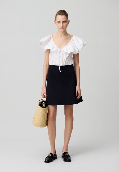 Blouse blanche à volants avec broderies florales, jupe noire évasée, mocassins noirs et sac en osier tressé avec des poignées noires.
