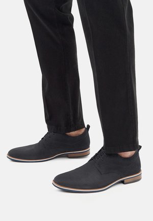 Schwarze, strukturierte Herrenschuhe mit runder Spitze, Schnürung und kontrastierender beiger Sohle. Kombiniert mit schwarzen Hosen.