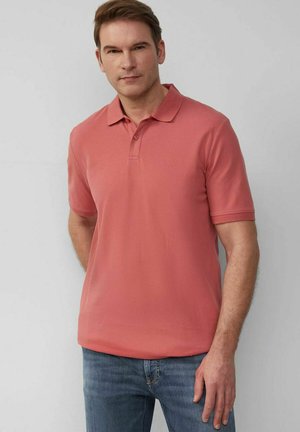 Mann trägt ein korallenpinke kurzärmelige Poloshirt und blaue Jeans, steht vor einem hellgrauen Hintergrund.