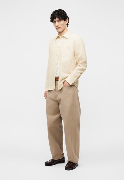 Νεαρός άνδρας όρθιος, φορεί κρεμ πουκάμισο με κουμπιά, μπεζ παντελόνι φαρδιάς γραμμής, καφέ ζώνη και σκούρα καφέ δερμάτινα παπούτσια σε λευκό φόντο.