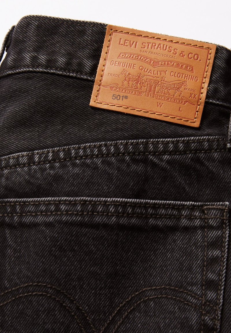 Jeans de mezclilla negra con un parche de cuero que presenta texto e imagen en relieve en la cinturilla. Costuras visibles en los bolsillos y costuras.