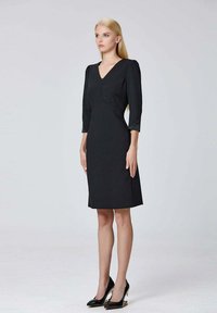 Robe noire avec un décolleté en V, des manches trois-quarts et des pinces de poitrine structurées. Tissu lisse et design jusqu'aux genoux. Portée avec des talons noirs.