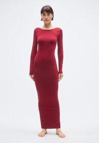 Šaty s dlhými rukávmi, priliehavé, vo výraznej bordovej farbe. Vyrobené z hladkej elastickej látky, majú lodičkový výstrih a dĺžku po členky. Modelka je bosá.