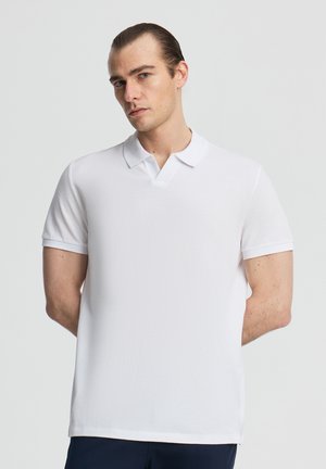 Młody mężczyzna z zaczesanymi do tyłu włosami, ubrany w prostą, białą koszulkę polo z krótkim rękawem, stojący z rękami za plecami na jasnym tle.