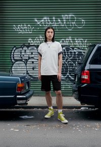 Világosszürke rövid ujjú ing felhajtott mandzsettákkal, fekete hálós rövidnadrág, limezöld tornacipő, két parkoló autó között állva, zöld graffiti előtt.