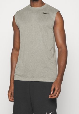 Man die een lichtgrijs mouwloos sportshirt en zwarte korte broek met Nike-logo's draagt, staande tegen een effen achtergrond.