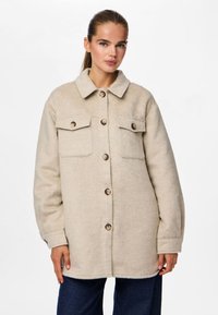 Beige skjortjacka i ullblandning med krage, fyra framfickor och stora bruna knappar. Har en avslappnad, oversize passform.