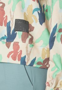 Pull avec imprimé floral coloré beige, comportant une étiquette noire avec le texte « Paul Smith » et des poignets élastiqués, accompagné d'un pantalon vert.