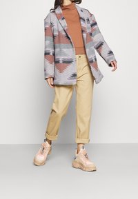 Blazer gris con motivos triangulares, suéter de cuello alto color óxido, pantalones beige con puños enrollados y zapatillas ligeras con detalles en rosa.