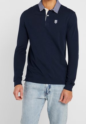 Homme portant un polo à manches longues bleu marine avec un col en chambray bleu et un jean délavé, debout contre un fond uni.