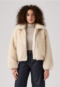 Cremegekleurde bomberjack van nepbont met een zachte textuur, een rits aan de voorkant en brede mouwen, gecombineerd met een beige turtleneck en donkere jeans.