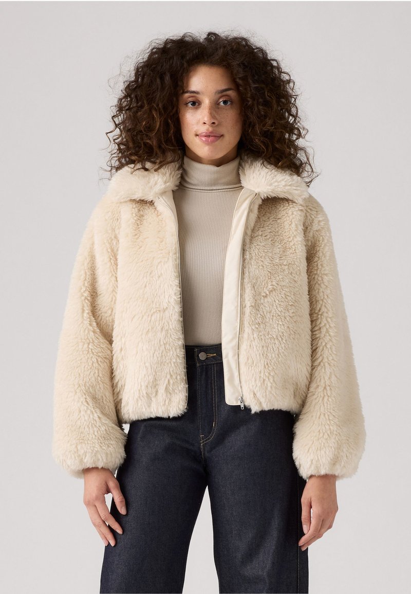 Veste bomber en fausse fourrure de couleur crème avec une texture douce, fermeture éclair sur le devant et manches larges, associée à un col roulé beige et un jean noir.