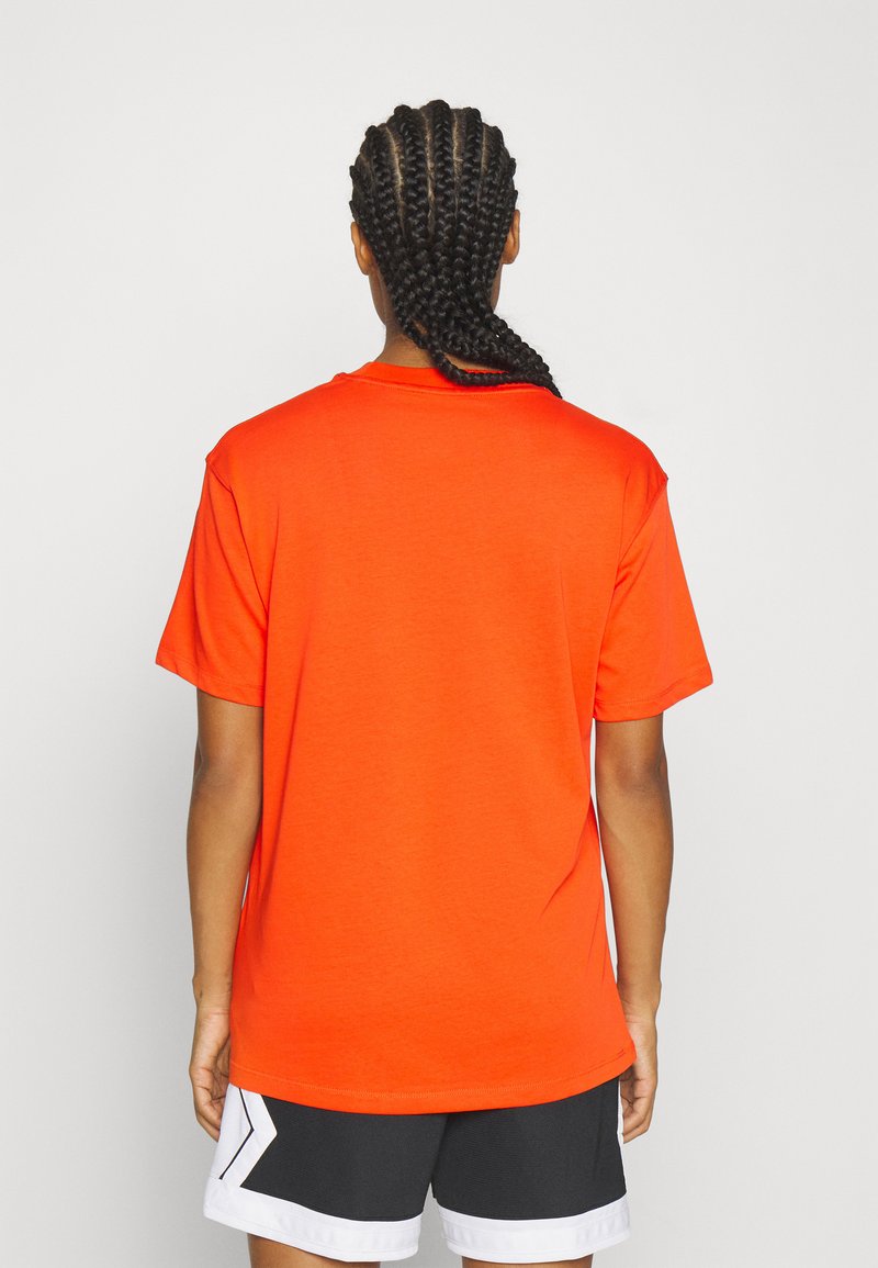 Personne avec de longs cheveux tressés portant une chemise à manches courtes orange vif et un short noir et blanc, de dos contre un arrière-plan uni.