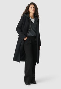 Vrouw met krullend haar die een lange zwarte jas draagt over een donker V-hals trui, grijze blouse, zwarte broek en zwarte puntige schoenen, staande tegen een grijze achtergrond.