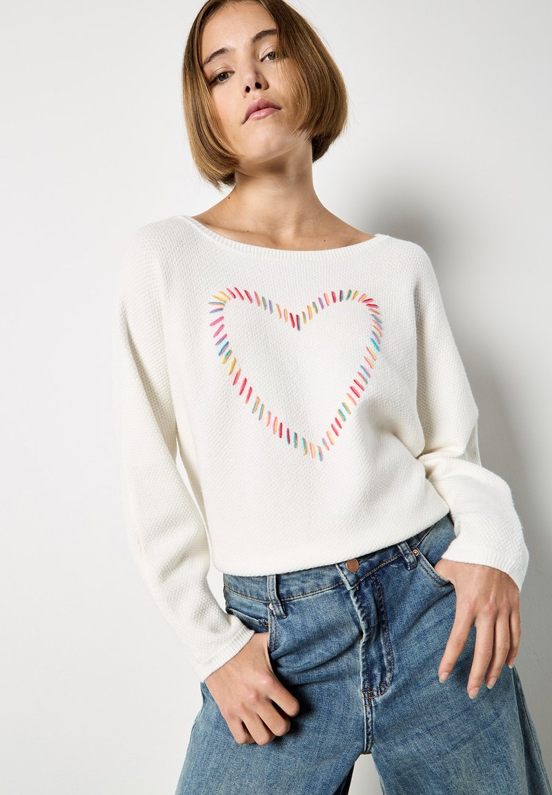 Pull blanc en tricot avec une broderie colorée en forme de cœur au centre. Encolure large et manches longues, associé à un jean en denim bleu.